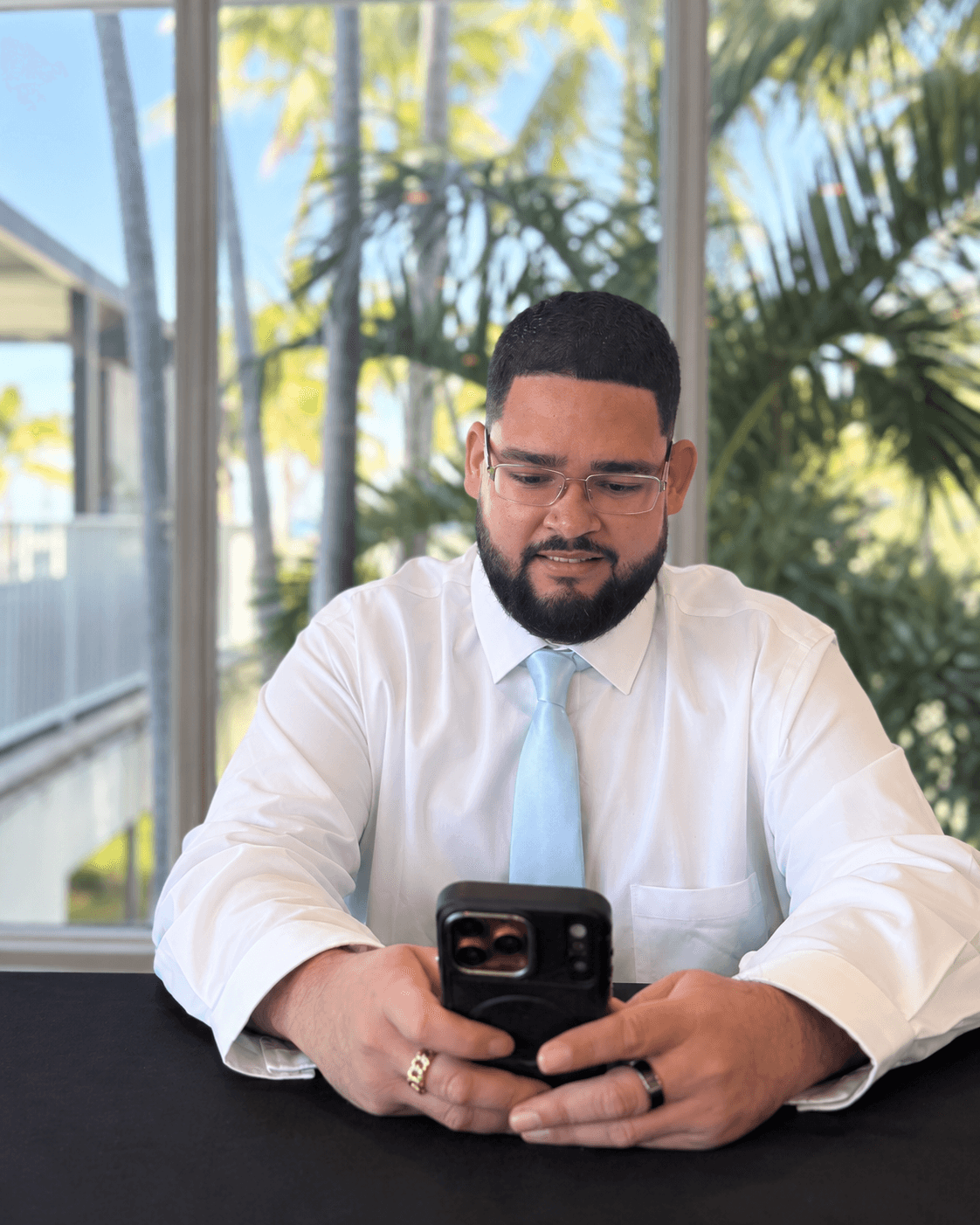 Emilio Sierra using his smartphone beside sunlit windows and tropical greenery.